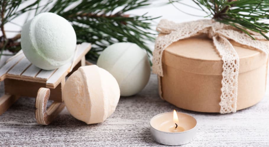 A relaxing scene with pastel-colored bath bombs, a wrapped gift box, and a lit tealight candle. The display is decorated with pine branches and a small wooden sled.