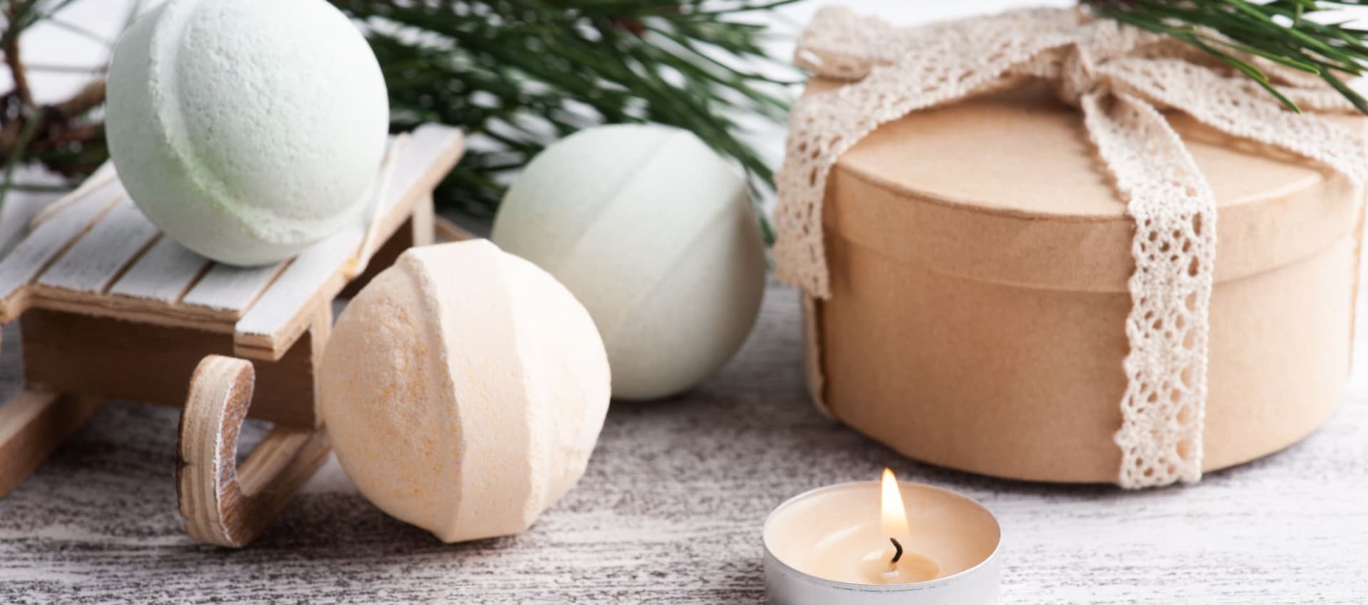 A relaxing scene with pastel-colored bath bombs, a wrapped gift box, and a lit tealight candle. The display is decorated with pine branches and a small wooden sled.