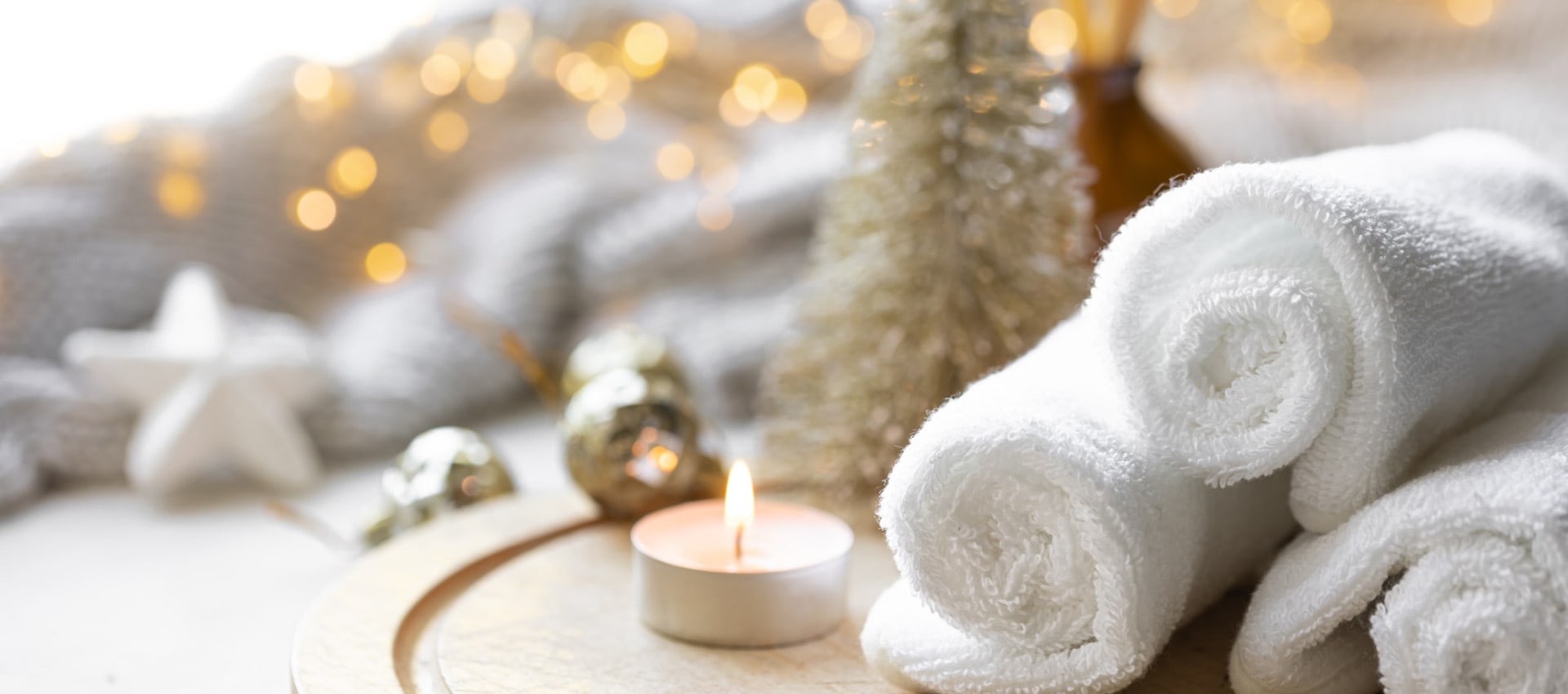 A serene spa-like setting with rolled white towels, a lit tealight candle, and festive decorations, including a small Christmas tree and twinkling lights in the background.