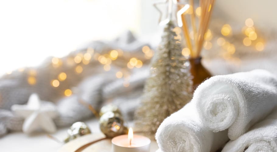 A serene spa-like setting with rolled white towels, a lit tealight candle, and festive decorations, including a small Christmas tree and twinkling lights in the background.