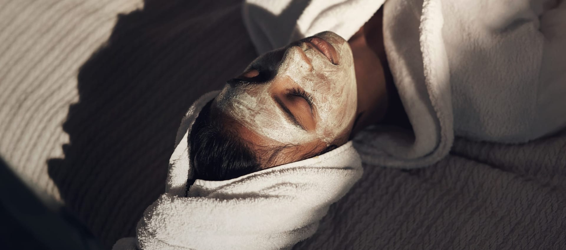 A person relaxing with a facial mask, wrapped in a plush white robe and towel. Soft sunlight casts shadows, creating a serene and spa-like atmosphere.
