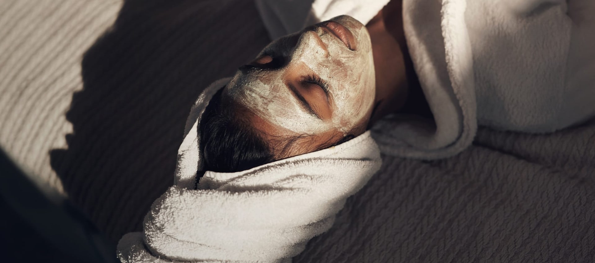 A person relaxing with a facial mask, wrapped in a plush white robe and towel. Soft sunlight casts shadows, creating a serene and spa-like atmosphere.