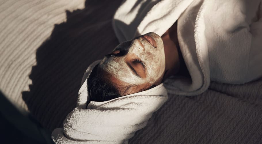 A person relaxing with a facial mask, wrapped in a plush white robe and towel. Soft sunlight casts shadows, creating a serene and spa-like atmosphere.