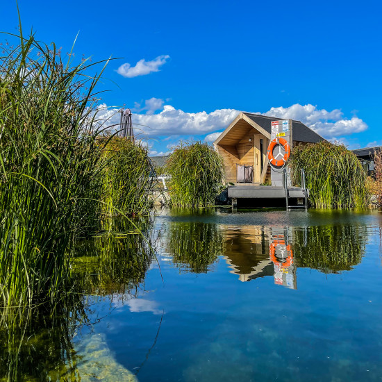 A finnish sauna sits on a clear blue natural swimming pond, with the sun shining.