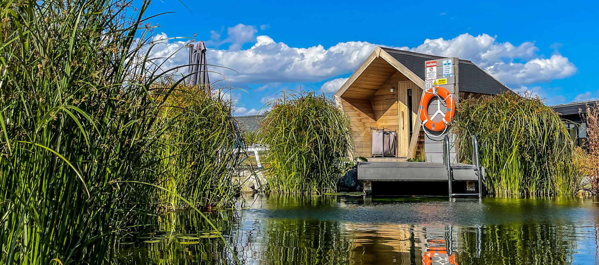 A finnish sauna sits on a clear blue natural swimming pond, with the sun shining.