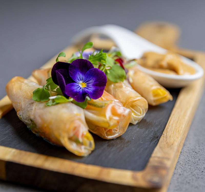 Fresh spring rolls served on a slate board with edible flowers, microgreens, and dipping sauce on a wooden tray for a refined dining presentation.