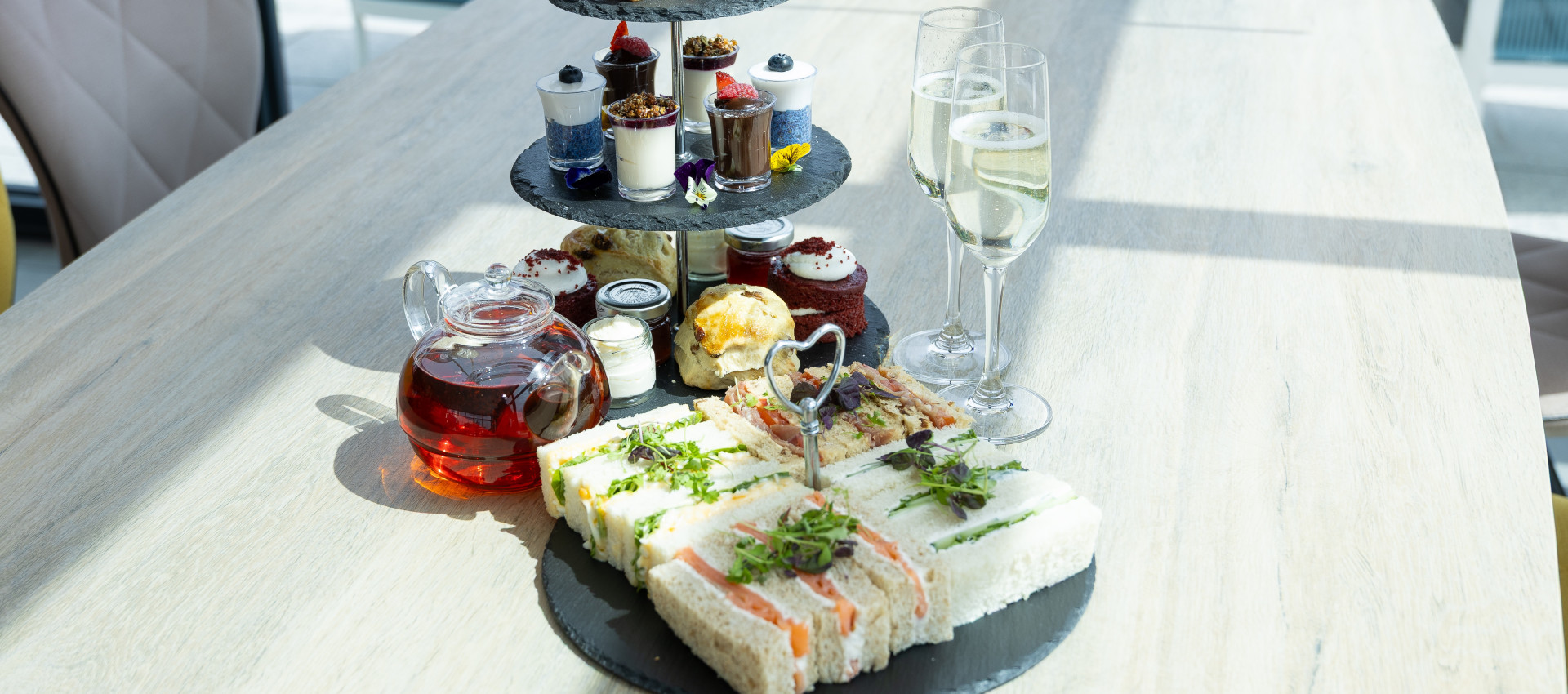 Afternoon tea stand with sandwiches, cakes, scones, and desserts, served with a teapot of tea and glasses of champagne on a light wooden table.