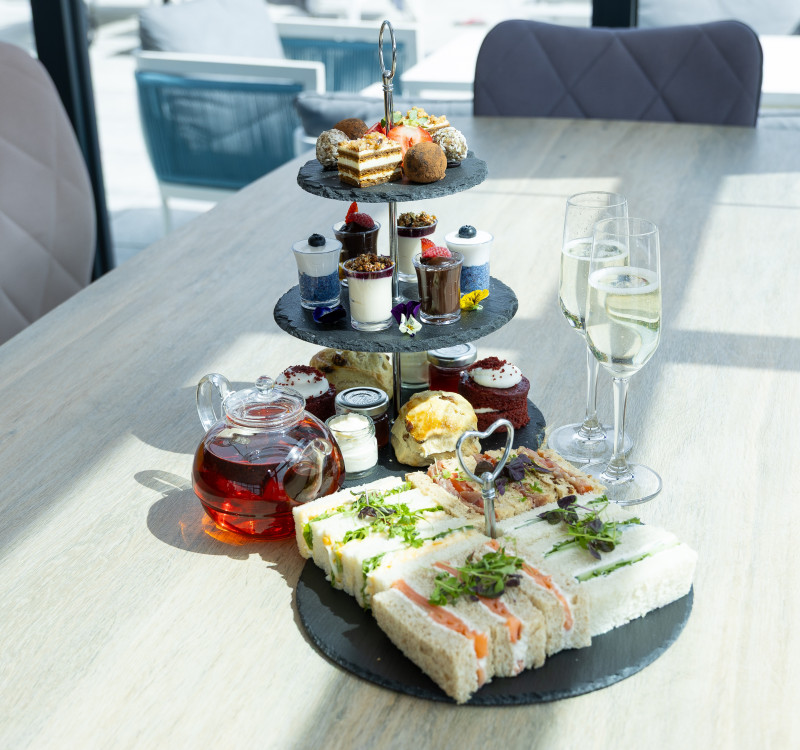 Afternoon tea stand with sandwiches, cakes, scones, and desserts, served with a teapot of tea and glasses of champagne on a light wooden table.