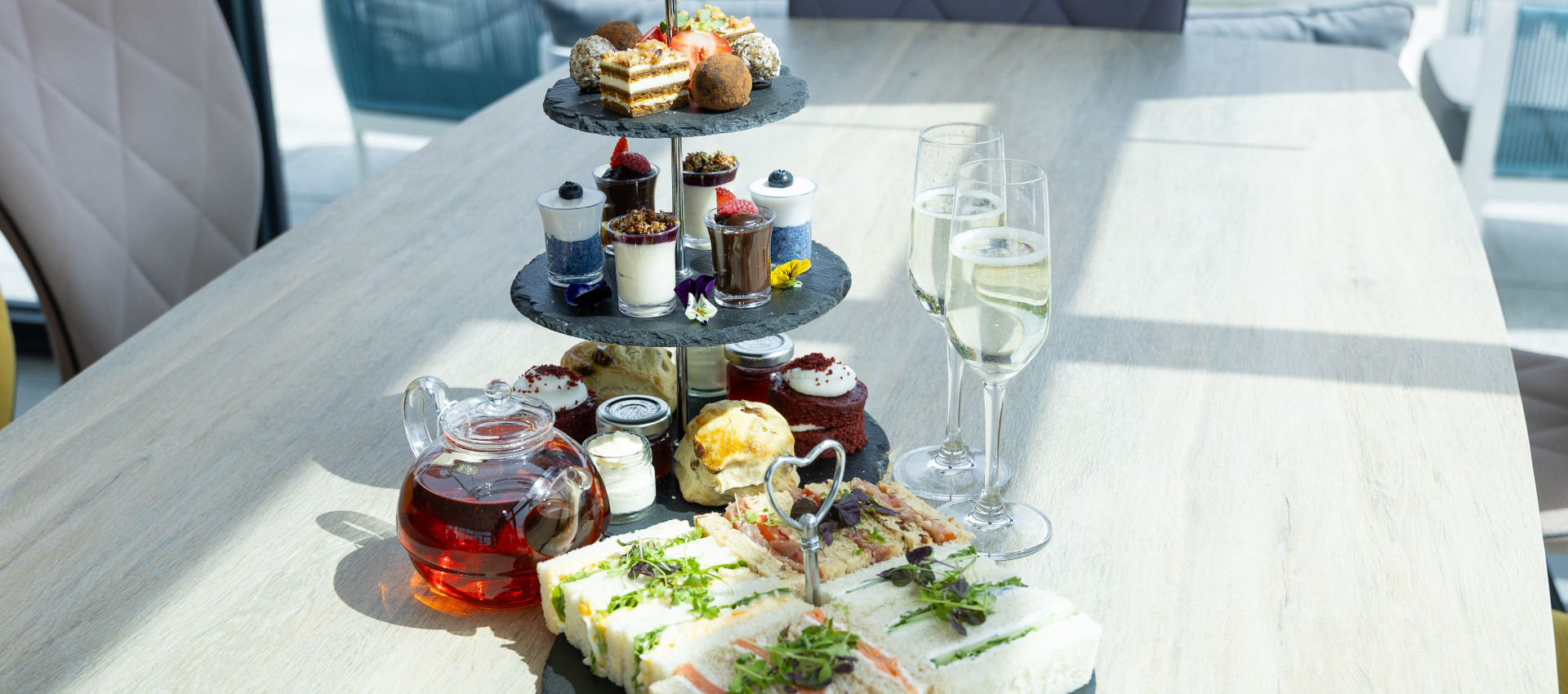 Afternoon tea stand with sandwiches, cakes, scones, and desserts, served with a teapot of tea and glasses of champagne on a light wooden table.
