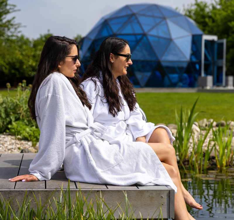 Two women in spa robes relax by a pond with their feet in the water at Glass House, with a blue wellness dome in the background.