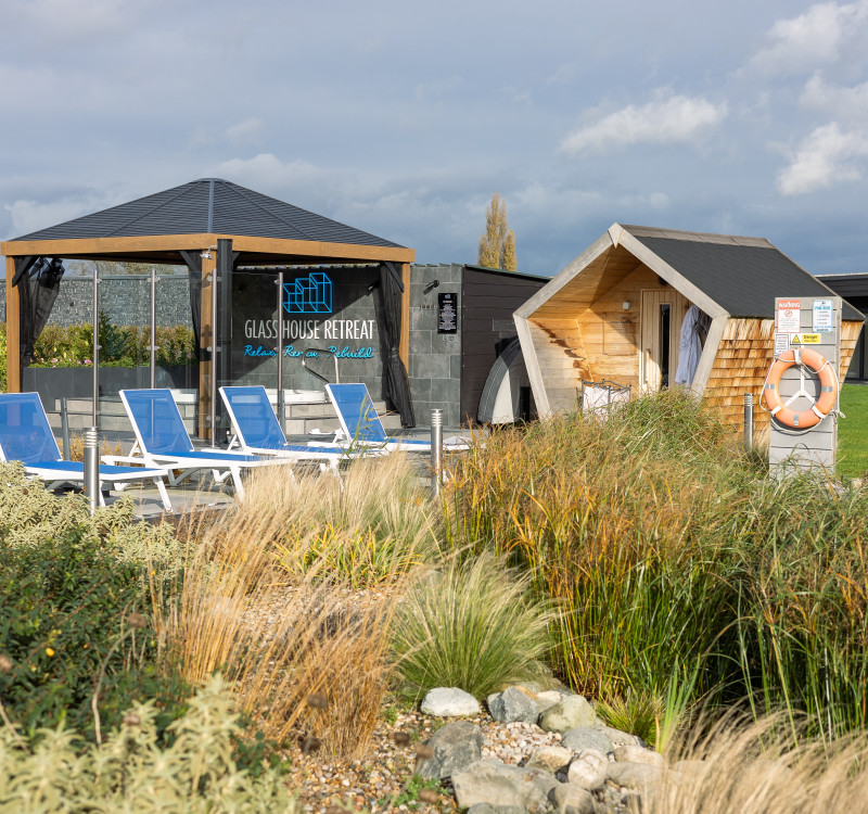 Outdoor spa area with sun loungers, modern wooden sauna hut, and a glass pavilion reading “Glass House Retreat,” surrounded by grasses and greenery.