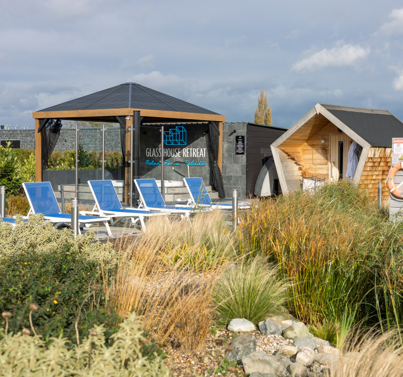 Outdoor spa area with sun loungers, modern wooden sauna hut, and a glass pavilion reading “Glass House Retreat,” surrounded by grasses and greenery.