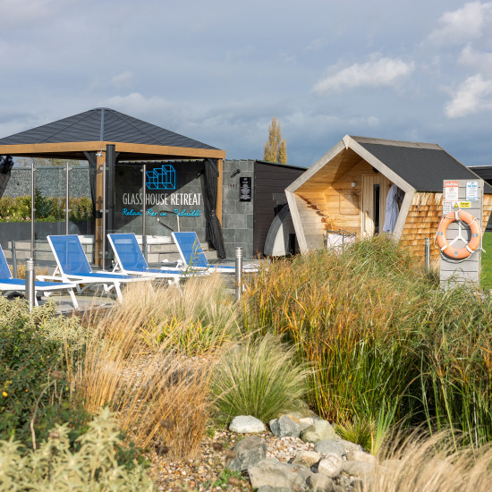 Outdoor spa area with sun loungers, modern wooden sauna hut, and a glass pavilion reading “Glass House Retreat,” surrounded by grasses and greenery.