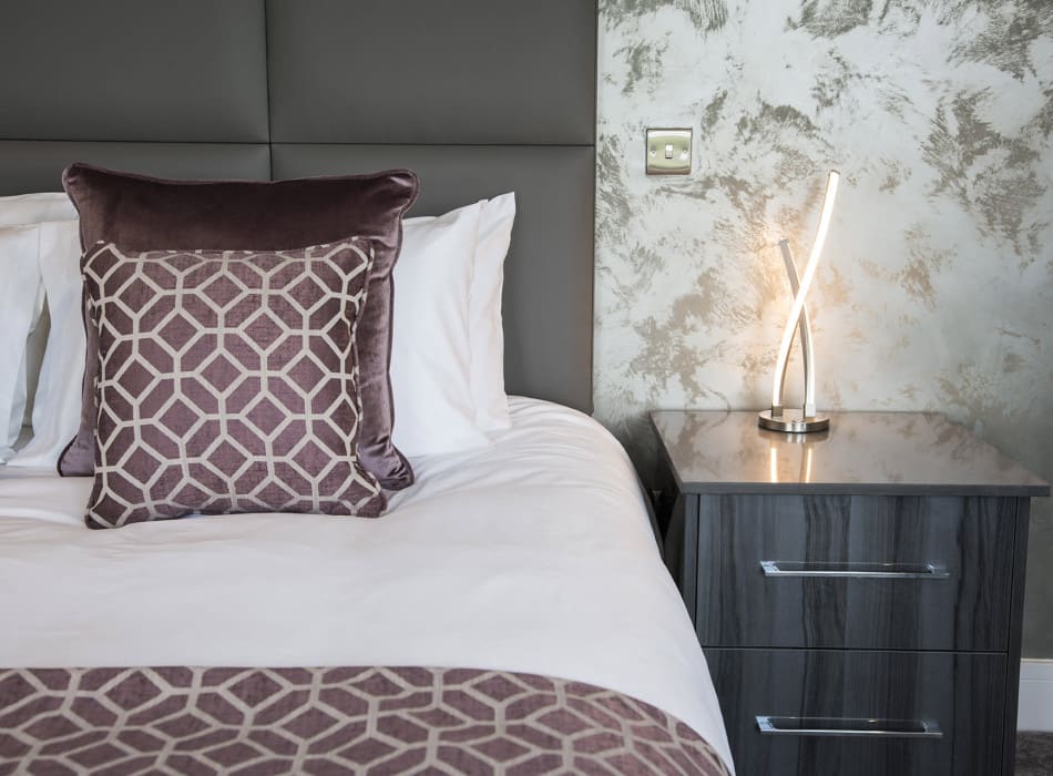 Bedside table with lamp next to cushions on bed